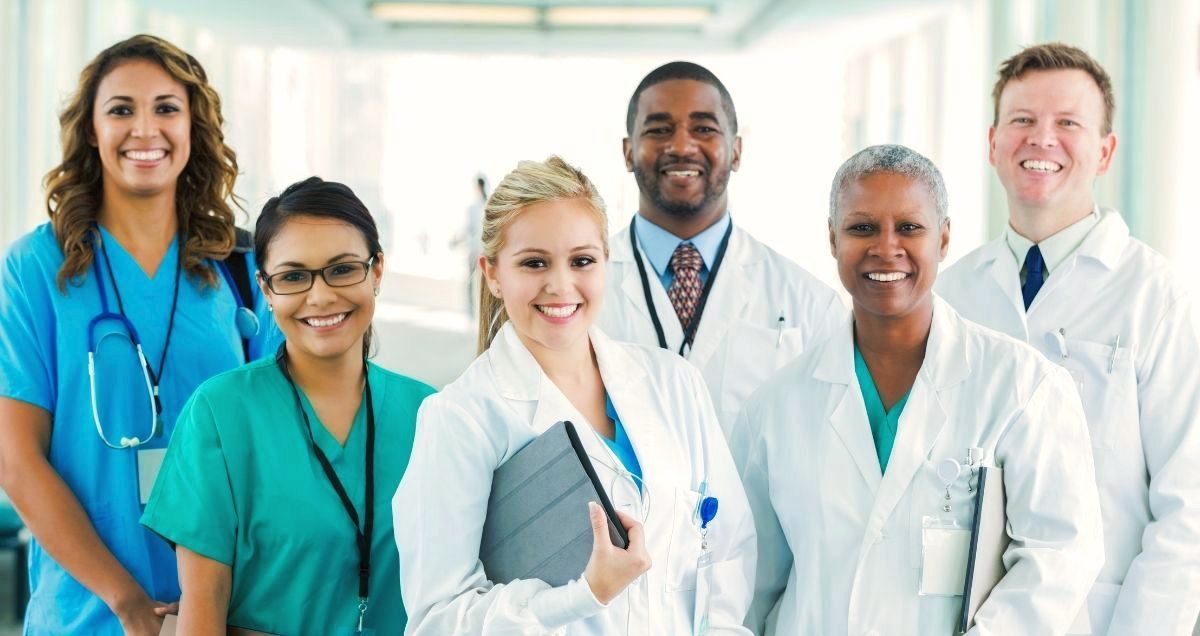 A diverse group of smiling doctors and nurses stand in a brightly lit hospital hallway.