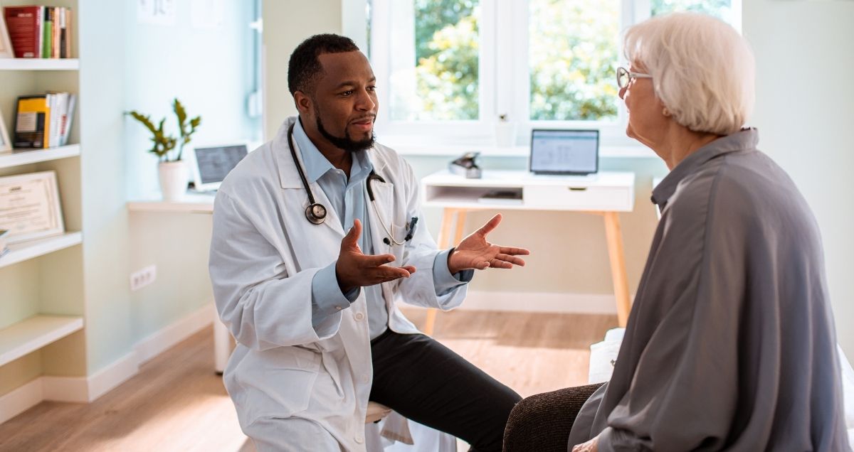 An African-American doctor with a lab coat and stethoscope talking to an elderly Caucasian patient in his office.