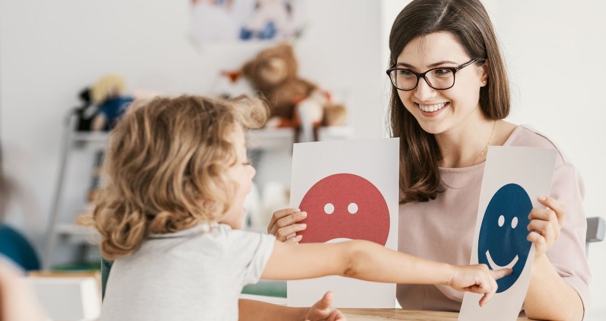 A child in a counseling session points to a sign with a smiling blue face that a therapist with glasses is holding.