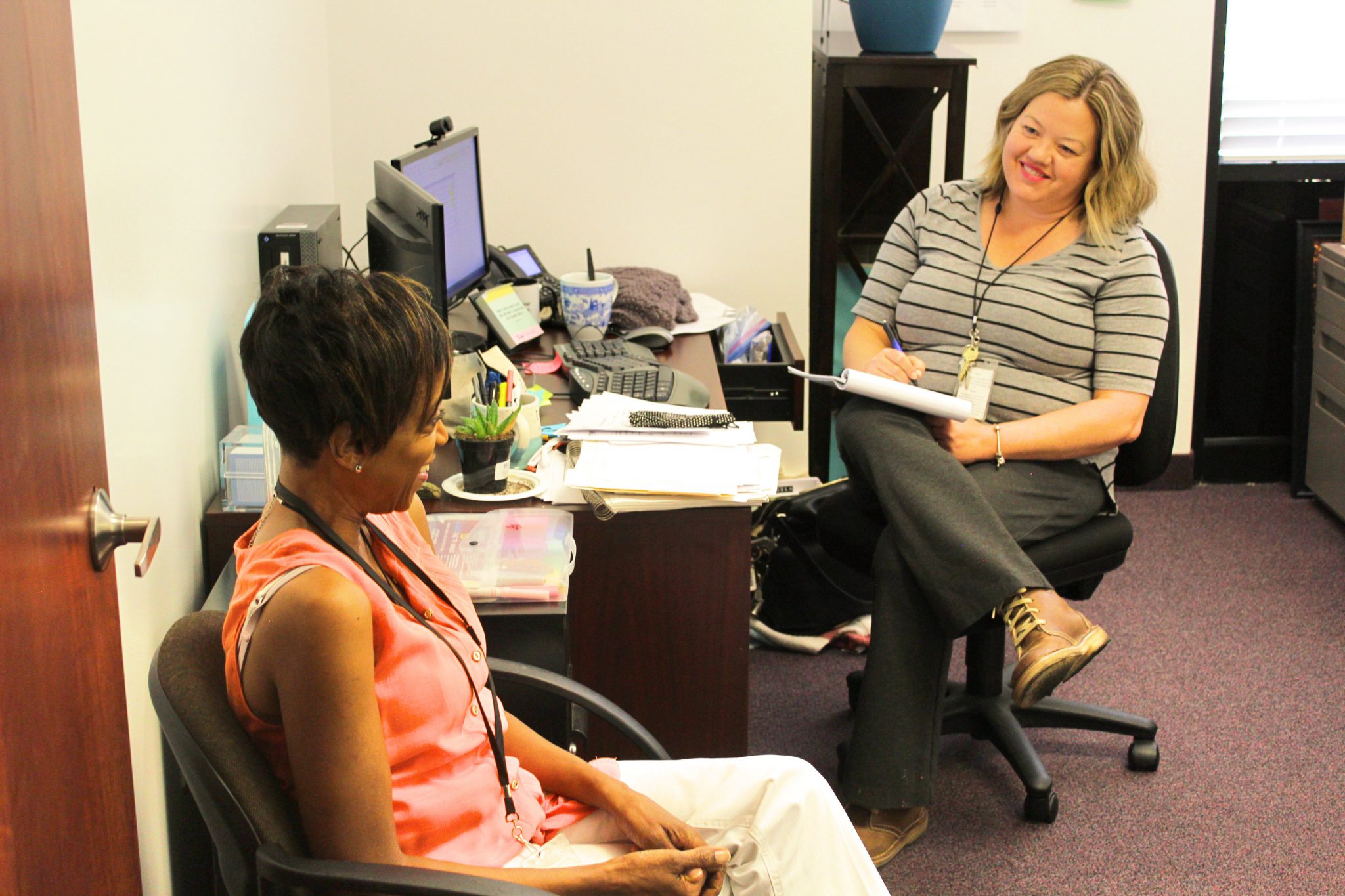 A smiling Psychiatrist takes notes on a notepad with a pen as she counsels her patient in her office.