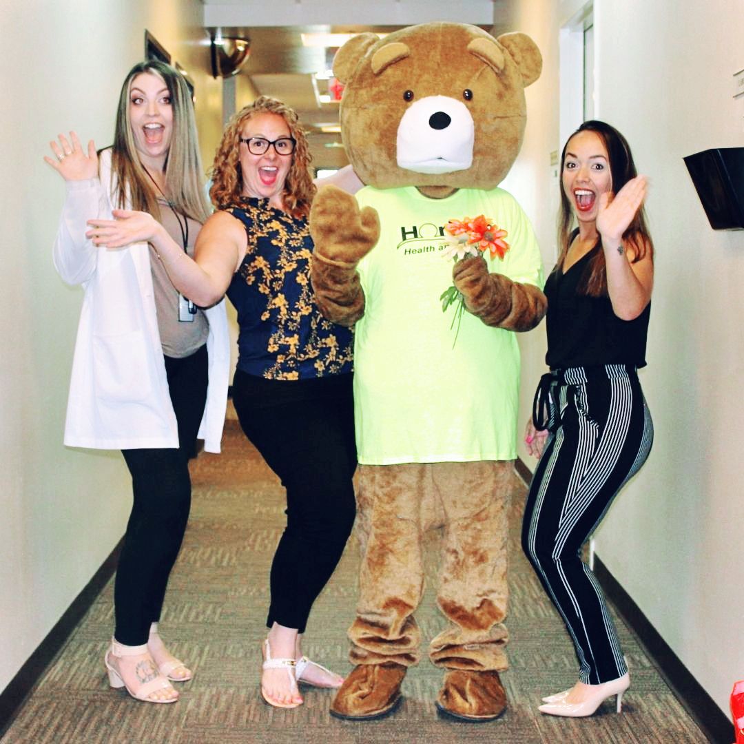 Cubby Bear with flowers and three happy staff members waving.