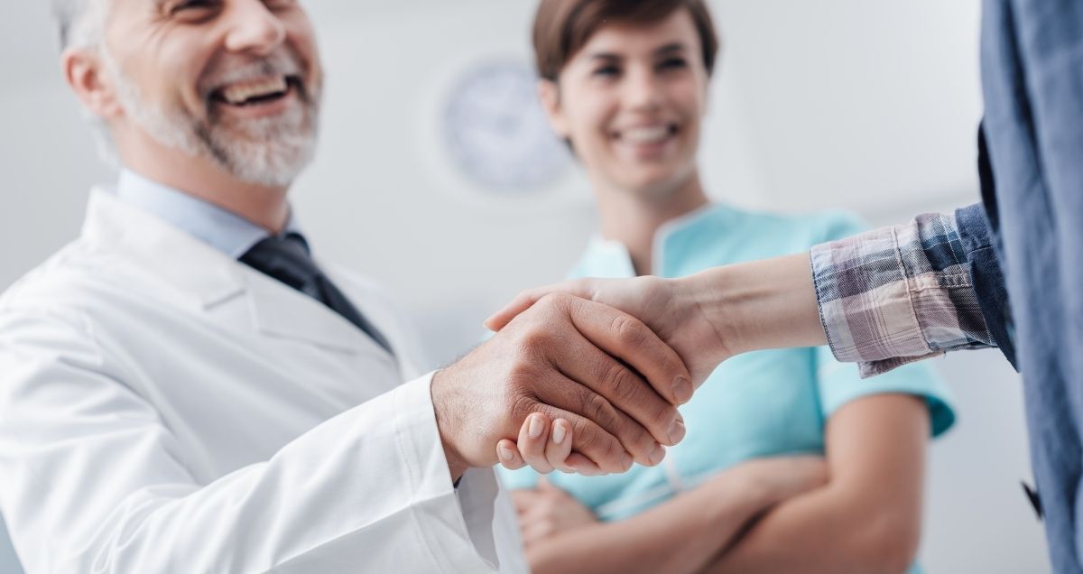 An elderly doctor shaking a patient's hand with a female nurse smiling in the background.