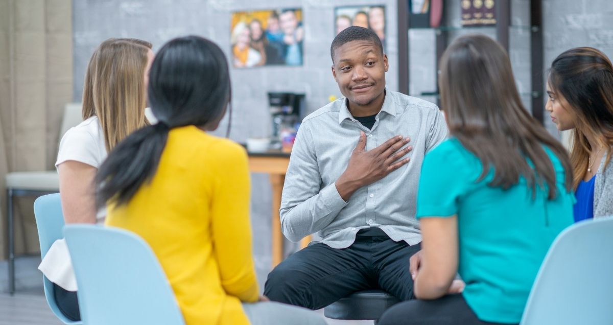 An emotional group therapy session with a diverse group of people listening to a man with his hand on his heart.