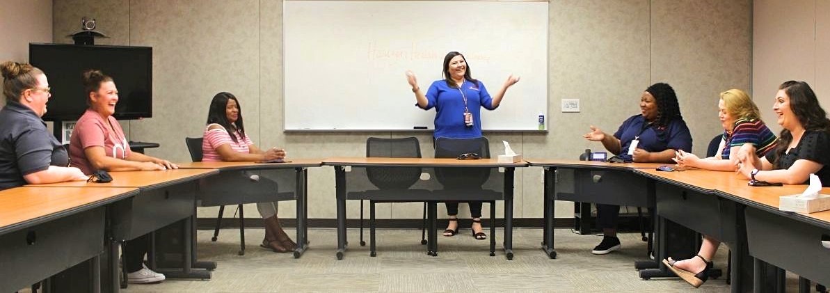 A group of laughing, female co-workers in a conference room with the standing presenter holding her hands up.