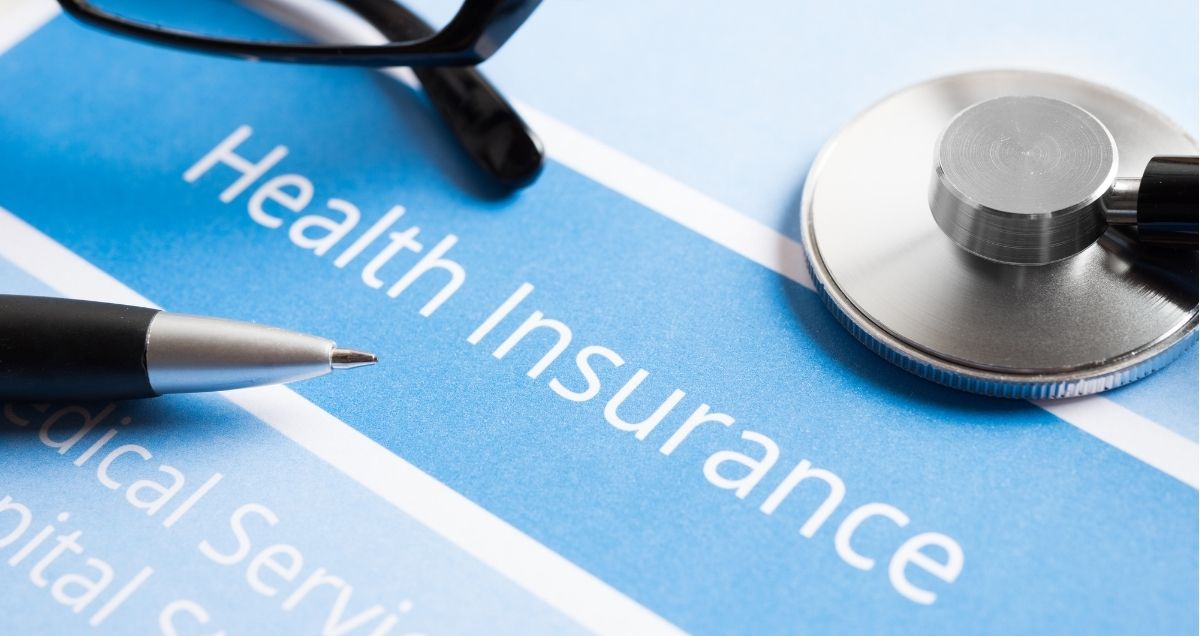 A pen, pair of glasses, and stethoscope are placed on top of a blue health insurance document.