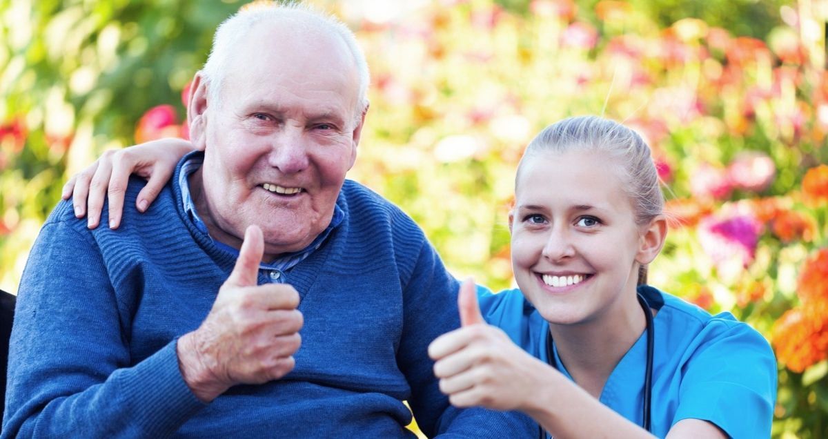 A smiling old man in a wheelchair and his kneeling nurse giving a thumbs up surrounded by tree leaves.