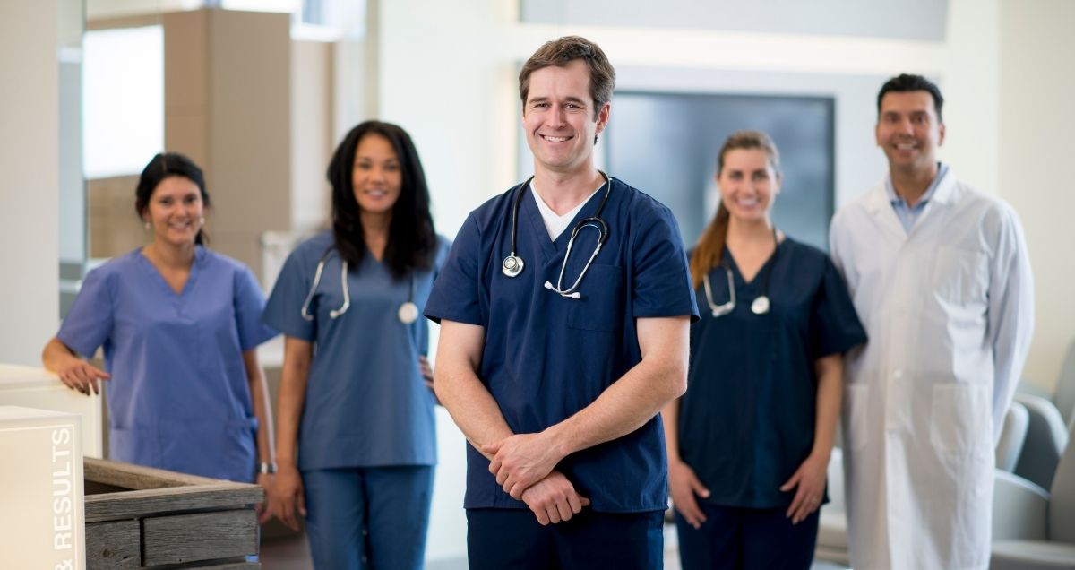 A middle-aged doctor standing in front of a diverse group of healthcare workers wearing scrubs and a lab coat.