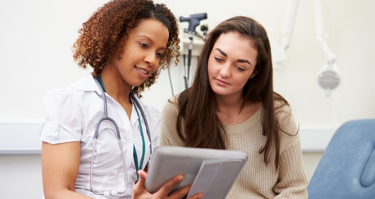 An African-American nurse with a stethoscope shows a Caucasian patient a clipboard as she sits in a doctor's office.