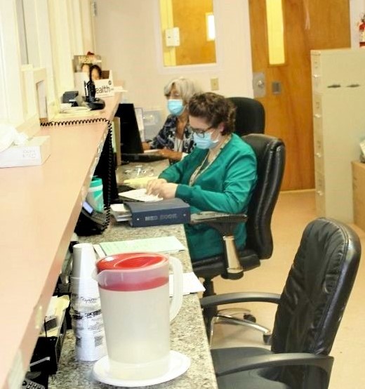 Female nurses in green scrubs wearing face masks are working at the desk of the in-patient treatment room.