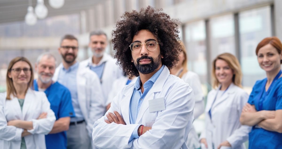 A confident African-American doctor wearing glasses standing in front of a group of medical staff.