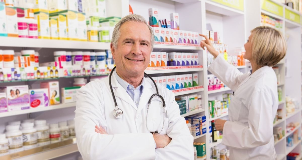 A pharmacist with arms crossed standing in front of shelves of medicine in a pharmacy.