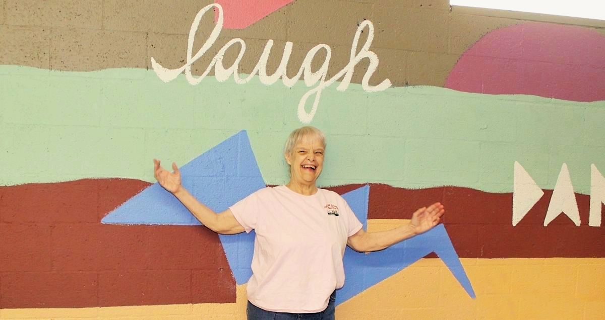A smiling disabled woman standing in front of a colorful wall with the word "laugh" painted on it.