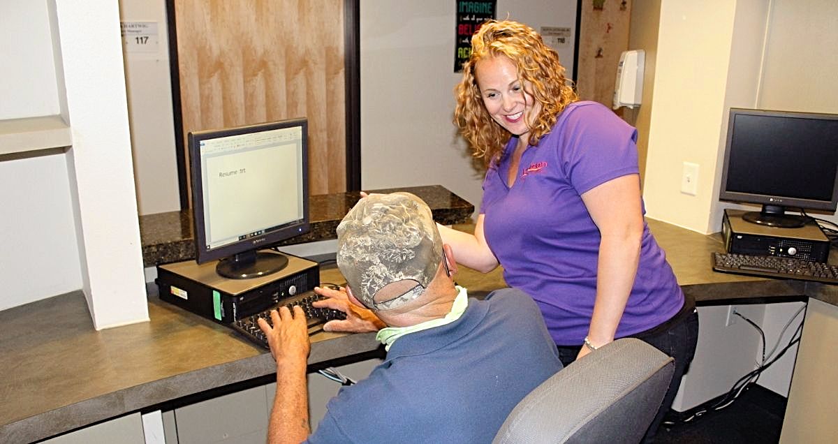 A smiling female employee helping a man type his resume on a computer in an office.