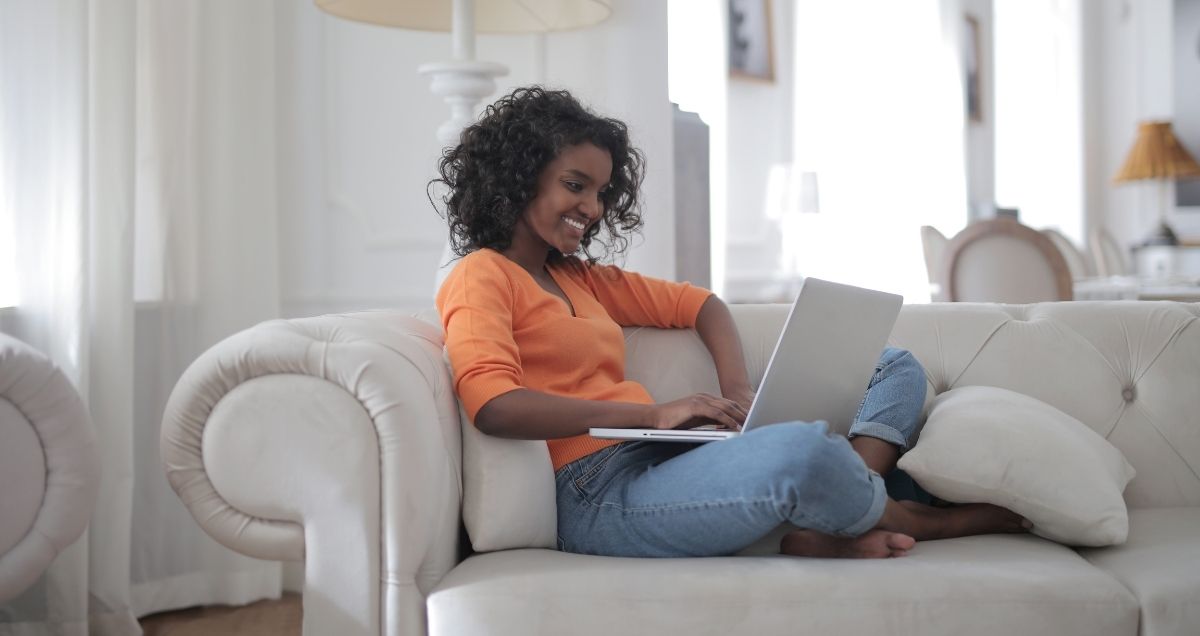 A smiling, African-American woman sitting on her couch in her home with a laptop on her lap.